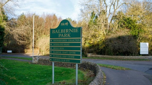A sign stating "Balbirnie Park" on a grass lawn next to a countryside road with trees along it.